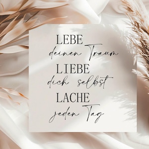 May include: A white card with black text that reads "LEBE deinen Traum LIEBE dich selbst LACHE jeden Tag". The card is on a white and beige background with dried pampas grass.