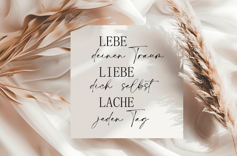 May include: A white card with black text that reads "LEBE deinen Traum LIEBE dich selbst LACHE jeden Tag". The card is on a white and beige background with dried pampas grass.