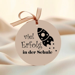May include: A round, beige gift tag with a brown ribbon. The tag features a black rocket ship graphic and the German text "viel Erfolg in der Schule," meaning "Good luck in school."