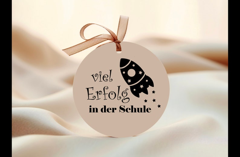 May include: A round, beige gift tag with a brown ribbon. The tag features a black rocket ship graphic and the German text "viel Erfolg in der Schule," meaning "Good luck in school."
