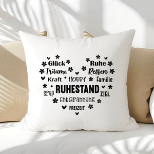 May include: White square pillow with a heart-shaped design filled with black text and floral accents. The text includes words like "Dreams", "Rest", and "Retirement". The pillow is resting on a beige sofa.
