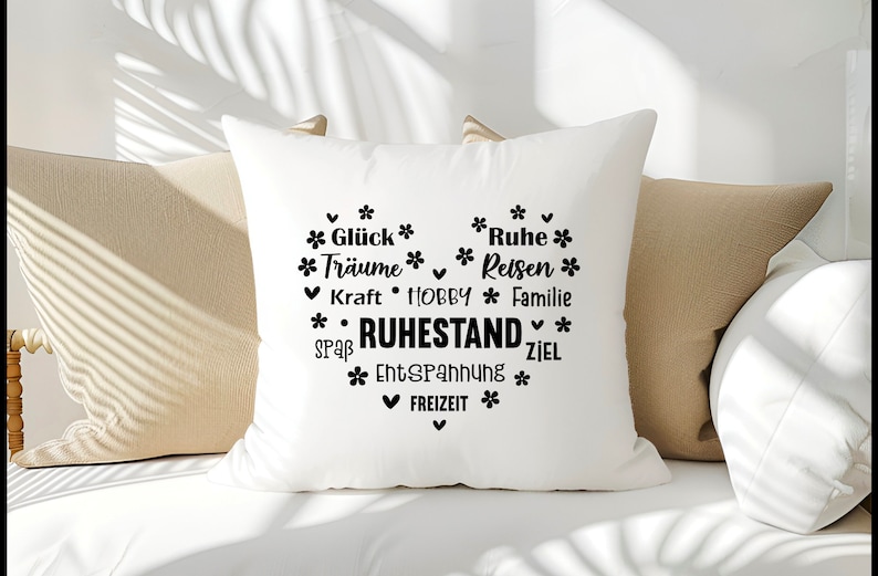May include: White square pillow with a heart-shaped design filled with black text and floral accents. The text includes words like "Dreams", "Rest", and "Retirement". The pillow is resting on a beige sofa.