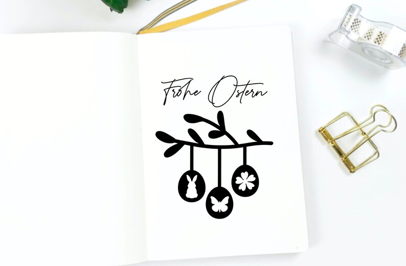 May include: A white notebook with the text "Frohe Ostern" written in black cursive. A black silhouette of a branch with three hanging Easter eggs is drawn below the text. The eggs are decorated with a bunny, a butterfly, and a flower.
