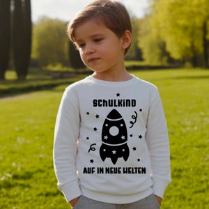 May include: A white long-sleeved shirt with a black rocket ship graphic and the text "Schulkind Auf in Neue Welten" printed on the front.