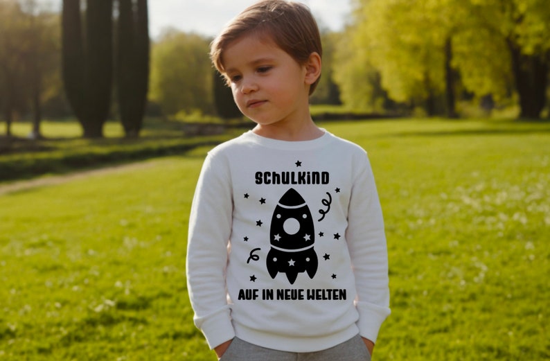 May include: A white long-sleeved shirt with a black rocket ship graphic and the text "Schulkind Auf in Neue Welten" printed on the front.