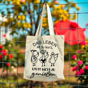 May include: A cream-coloured tote bag featuring the German phrase "DAS LEBEN ist zu kurz, UM ES NICHT zu genie&szlig;en." Below the text are three cartoon birds, each with a glass. The bag is suspended from a wire fence, with flowers in the background.