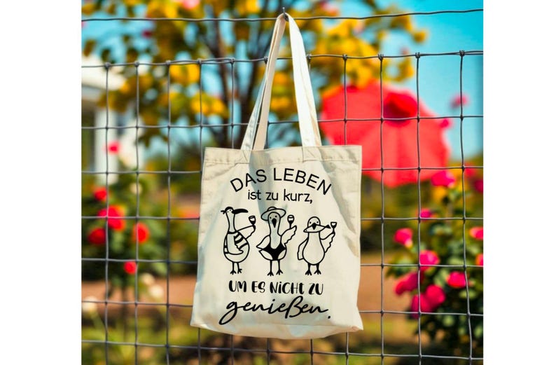 May include: A cream-coloured tote bag featuring the German phrase "DAS LEBEN ist zu kurz, UM ES NICHT zu genie&szlig;en." Below the text are three cartoon birds, each with a glass. The bag is suspended from a wire fence, with flowers in the background.