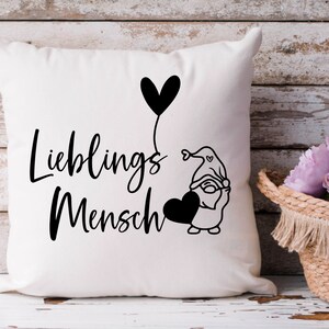 May include: White cushion with black text that reads "Lieblings Mensch" and a black and white illustration of a gnome holding a heart-shaped balloon.