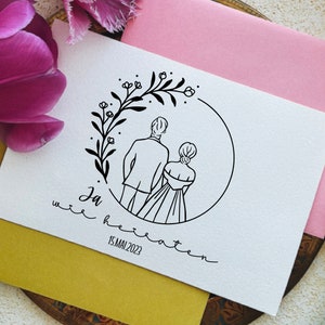 May include: A white card with a black line drawing of a couple standing back to back in a circle of flowers. The text "Ja  wir heiraten 15.MAI 2023" is written below the drawing.