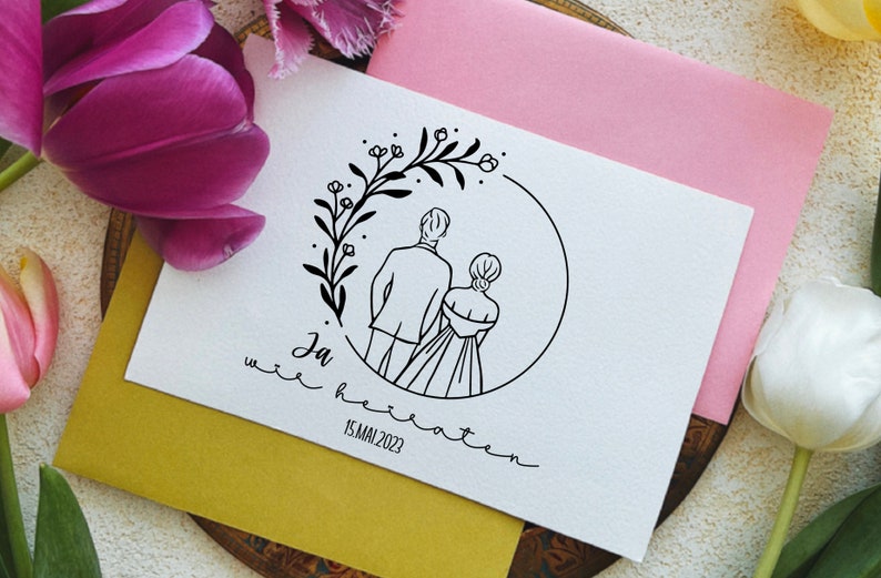 May include: A white card with a black line drawing of a couple standing back to back in a circle of flowers. The text "Ja  wir heiraten 15.MAI 2023" is written below the drawing.