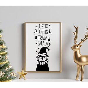 May include: A black and white printable art print with a cat wearing a Santa hat and the text "Lustig Lustig Trala Lalala". The print is framed in a gold frame and is displayed on a shelf with a gold reindeer figurine and a small Christmas tree.