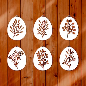 May include: Ten white wooden Easter egg ornaments with different floral designs cut out. The ornaments are hanging on a wood panel background.