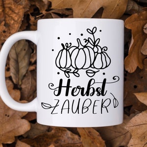 May include: White ceramic mug with a black outline of pumpkins and leaves. The text "Herbst Zauber" is written in black cursive.