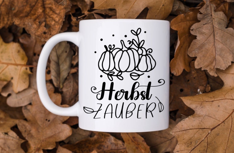 May include: White ceramic mug with a black outline of pumpkins and leaves. The text "Herbst Zauber" is written in black cursive.