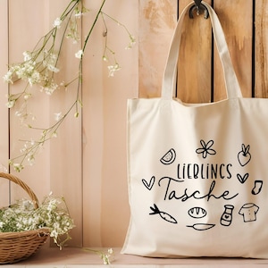 May include: A white canvas tote bag with black text and illustrations. The text reads "Lieblings Tasche" which translates to "Favorite Bag". The illustrations include a watermelon slice, a flower, a carrot, a loaf of bread, a bottle of sauce, a t-shirt, and a heart.