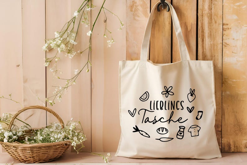 May include: A white canvas tote bag with black text and illustrations. The text reads "Lieblings Tasche" which translates to "Favorite Bag". The illustrations include a watermelon slice, a flower, a carrot, a loaf of bread, a bottle of sauce, a t-shirt, and a heart.
