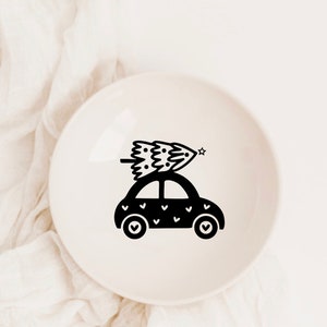 May include: A black and white illustration of a car with a Christmas tree on top. The car is decorated with small hearts. The illustration is on a white ceramic plate.