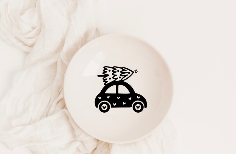 May include: A black and white illustration of a car with a Christmas tree on top. The car is decorated with small hearts. The illustration is on a white ceramic plate.