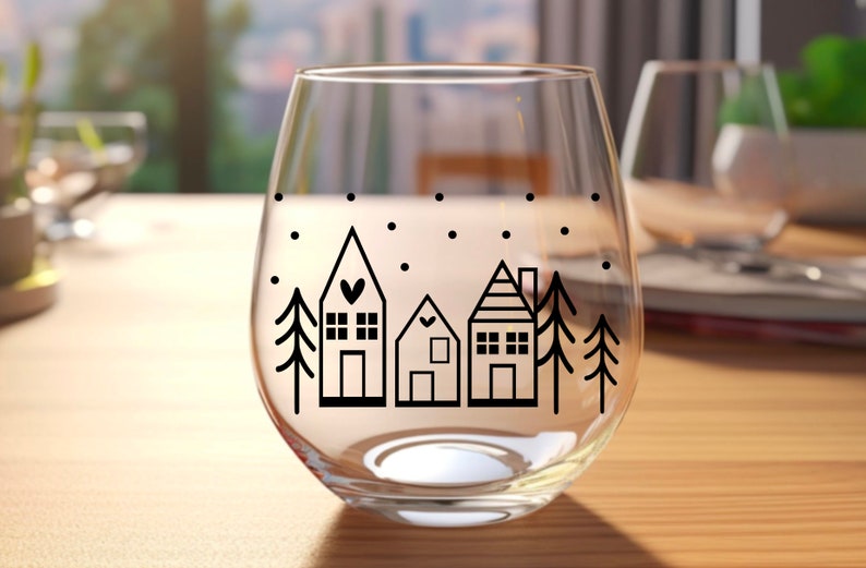 May include: A clear glass wine glass with a black and white design of a snowy village with three houses and two trees. The houses have small windows and doors, and the trees are simple silhouettes.