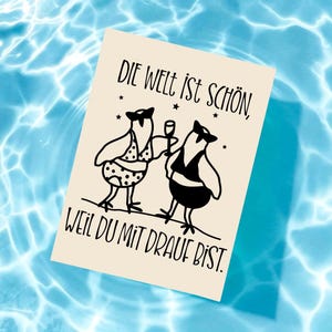May include: A beige card with black text and illustrations floats on blue water. The card features two cartoon birds, one in a polka-dot swimsuit and the other in a tuxedo, with the German phrase "Die Welt ist sch&ouml;n, weil du mit drauf bist."
