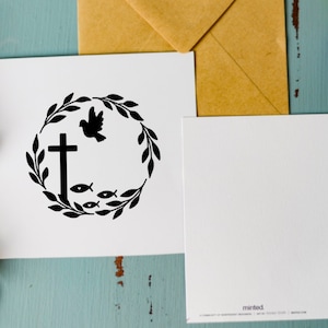 May include: A white card with a black and white illustration of a cross, a dove, and fish inside a wreath. The card is on a blue wooden surface with a brown envelope and purple flowers.