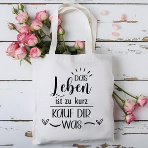 May include: White tote bag with black text that reads "Das Leben ist zu kurz, kauf dir was" with two hearts on either side of the text. The bag is surrounded by pink roses.