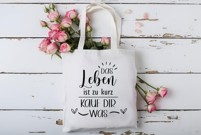 May include: White tote bag with black text that reads "Das Leben ist zu kurz, kauf dir was" with two hearts on either side of the text. The bag is surrounded by pink roses.