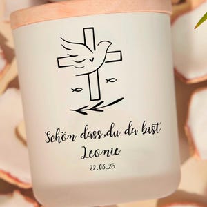 Pu&ograve; includere: Candela bianca con un coperchio in legno. La candela presenta un design in bianco e nero di una colomba con le ali spiegate, una croce e due simboli di pesci. Il testo "Sch&ouml;n dass du da bist Leonie 22.03.25" &egrave; stampato sotto il design.