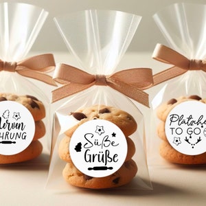 May include: Three clear cellophane bags tied with brown ribbon, each containing a stack of three chocolate chip cookies with a round sticker on top. The stickers have different text in black lettering: "Nerven Nahrung", "S&uuml;&szlig;e Gr&uuml;&szlig;e", and "Pl&auml;tzchen TO GO".