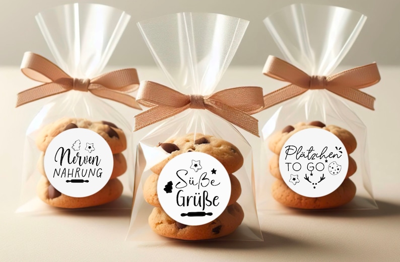 May include: Three clear cellophane bags tied with brown ribbon, each containing a stack of three chocolate chip cookies with a round sticker on top. The stickers have different text in black lettering: "Nerven Nahrung", "S&uuml;&szlig;e Gr&uuml;&szlig;e", and "Pl&auml;tzchen TO GO".