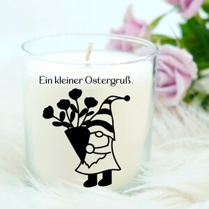 May include: A white candle in a glass jar with a black silhouette of a gnome holding a bouquet of flowers. The text "Ein kleiner Ostergru&szlig;." is printed on the jar.