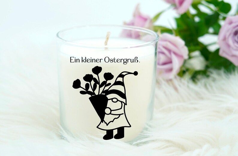 May include: A white candle in a glass jar with a black silhouette of a gnome holding a bouquet of flowers. The text "Ein kleiner Ostergru&szlig;." is printed on the jar.