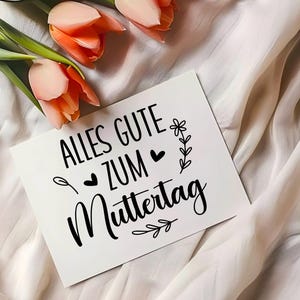 May include: A white card with black text that reads "Alles Gute zum Muttertag" with a small flower and heart design. The card is on a white fabric background with pink flowers.