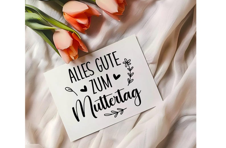 May include: A white card with black text that reads "Alles Gute zum Muttertag" with a small flower and heart design. The card is on a white fabric background with pink flowers.