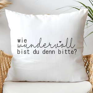 May include: White throw pillow with black text that reads "wie wundervoll bist du denn bitte?"  The pillow is sitting on a wicker chair.