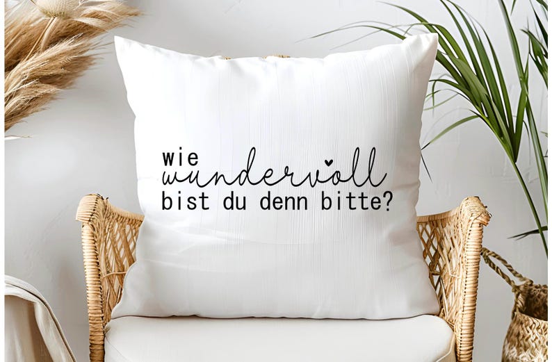 May include: White throw pillow with black text that reads "wie wundervoll bist du denn bitte?"  The pillow is sitting on a wicker chair.