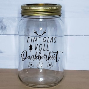 May include: A clear glass jar with a gold lid and a black vinyl sticker that reads "Ein Glas Voll Dankbarkeit" with a small tree and star design.