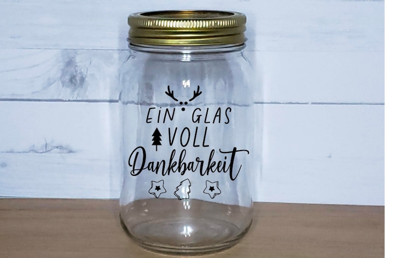 May include: A clear glass jar with a gold lid and a black vinyl sticker that reads "Ein Glas Voll Dankbarkeit" with a small tree and star design.