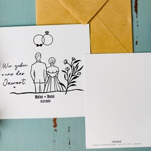 May include: A white card with a black line drawing of a couple getting married. The text on the card reads "Wir geben uns das Jawort" and "Mateo & Annie 13.07.2023".