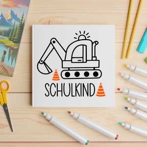 May include: A white notebook with a black and white illustration of an excavator with orange traffic cones. The text "SCHULKIND" is written below the excavator.