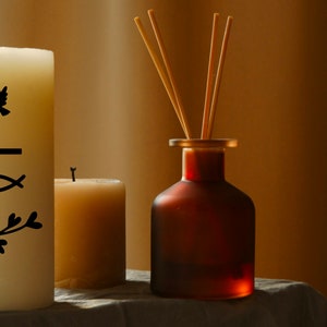 May include: Two white candles, one with a black design of a dove, cross, fish, and branches, and the other plain. A brown glass bottle with wooden sticks for a diffuser.