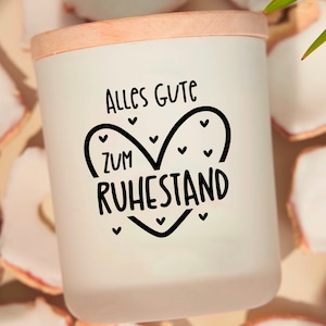 May include: A white candle holder with a wooden lid. The candle holder has the words "Alles Gute zum Ruhestand" in black script inside a heart shape. The background has white and tan decorative pieces.