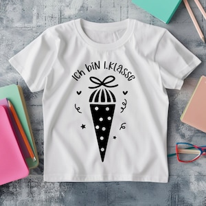 May include: White t-shirt with a black graphic of a school cone and the text "Ich bin 1. Klasse". The design includes hearts, stars, and swirls. The shirt is on a gray surface with school supplies.