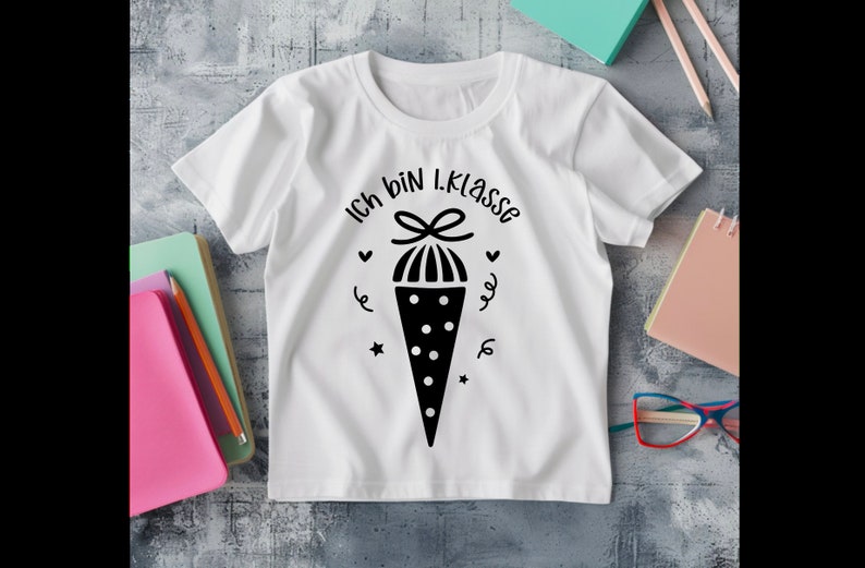 May include: White t-shirt with a black graphic of a school cone and the text "Ich bin 1. Klasse". The design includes hearts, stars, and swirls. The shirt is on a gray surface with school supplies.