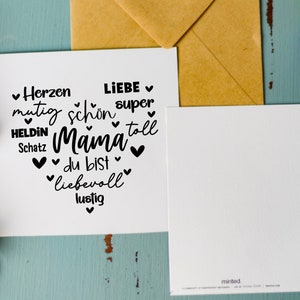Może przedstawiać: Biała kartka z czarnym tekstem, kt&oacute;ry brzmi "Herzen mutig Heldin Schatz Mama du bist liebevoll lustig Liebe super toll" z sercami wok&oacute;ł tekstu. Kartka leży na niebieskiej powierzchni z brązową kopertą i fioletowymi kwiatami.