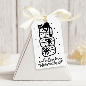 May include: A white gift tag with a black and white illustration of a cat sitting on top of a stack of presents. The text on the tag reads "erholsame Weihnachtszeit".