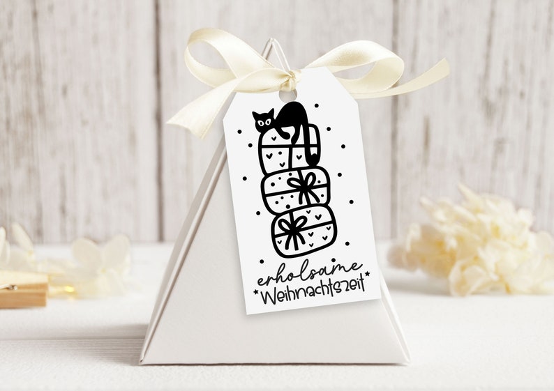 May include: A white gift tag with a black and white illustration of a cat sitting on top of a stack of presents. The text on the tag reads "erholsame Weihnachtszeit".