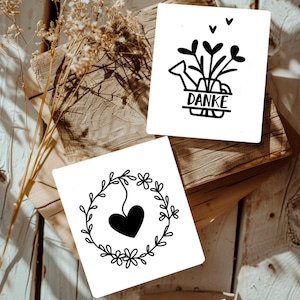May include: Two white cards with black illustrations. The first card has a black watering can with flowers and the word "DANKE" written on a banner. The second card has a black heart inside a wreath of leaves and flowers.