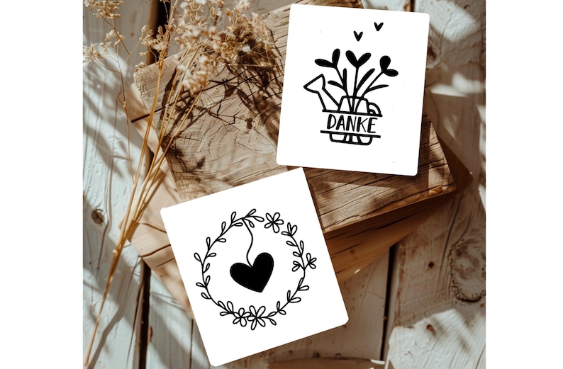 May include: Two white cards with black illustrations. The first card has a black watering can with flowers and the word "DANKE" written on a banner. The second card has a black heart inside a wreath of leaves and flowers.
