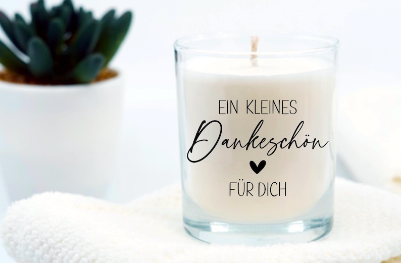 May include: A clear glass candle jar with a white candle inside. The candle has a black label with the text "Ein kleines Dankesch&ouml;n f&uuml;r dich" and a small black heart.